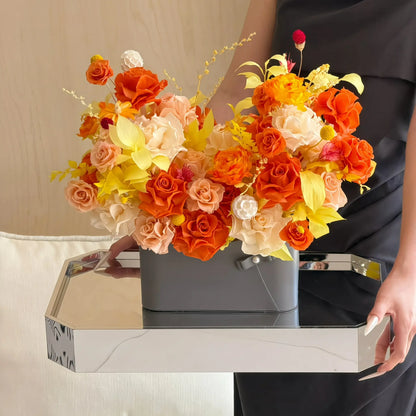 LULU ROSÉE Sunset Orange Parisian Bloom Garden Preserved Flower Arrangement in a gray vase on a table with a person partially visible