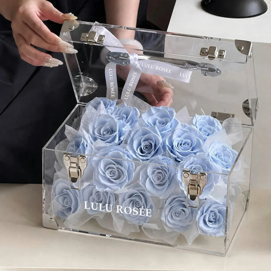 LULU ROSÉE Petite Eternal Acrylic Handbag Rose Box, small transparent acrylic handbag-shaped gift box filled with soft baby blue preserved roses with open lid, handheld lifestyle shot, customizable flower color options, mini luxury everlasting floral souvenir for birthday, anniversary & romantic gift giving